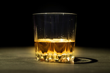 A glass of whiskey lit by a ray of light stands on a dark table and on a black background