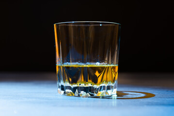 A glass of whiskey lit by a ray of light stands on a dark table and on a black background