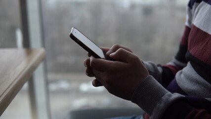 A young man with a phone closeup. A man sits in a cafe by the panoramic window with a smartphone in his hands. View from the window.