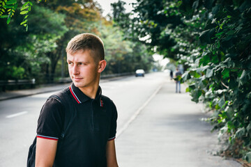 Portrait of a young man in a black T-shirt looking back on the background of a road and green trees.