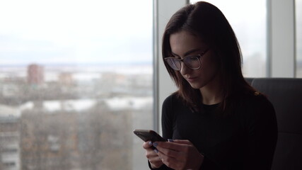 A young woman with a phone. A girl sits in a cafe by the panoramic window with a smartphone in her hands. View from the window.