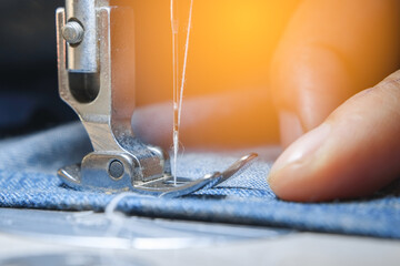 Macro shoot hand of the seamstress is using industrial sewing machine to sew the seams of blue jeans with orange light close-up.