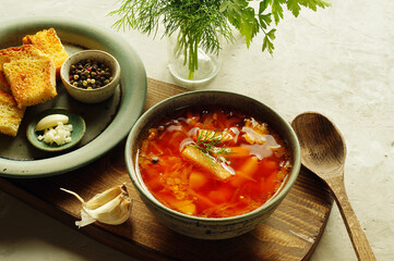 A bowl of soup-borscht, top decorated with a sprig of dill, close-up, on a wooden Board with a spoon.Next to it on a plate is toasted, crunchy bread with garlic and a sauce pot with peppers. 