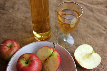 Homemade apple cider in a bottle and in a glass, and fresh apples on a burlap background, top view, copy space. Red apples in a ceramic bowl