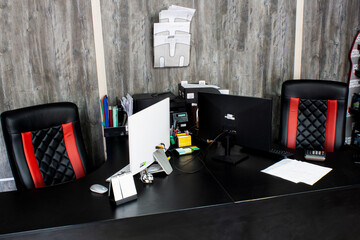 Workspace in office with computers. Monitors on black tables with director's leather chairs. Paper, documents, keyboard, mouse, calculator, printer, stickers, pens, pencils on wooden wall background