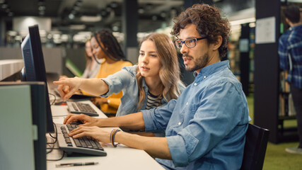 University Library: Bright Caucasian Girl and Smart Boy Together Work on Computers, Chat, Discuss Class Assignment, Explain and Advice Each Other. Diverse Group of Students Exam Study Teamwork
