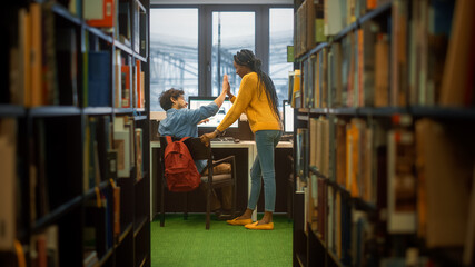 University Library: Boy Uses Personal Computer at His Desk, Talks with Girl Classmate Give High Five after Successful Solution. Focused Students Study Together. Shot Between Rows of Bookshelves