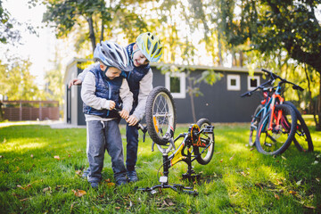 Two children, older boys and younger brother learning repair bike. Two guys siblings in helmets and single clothes use pump tool and pump air into bicycle wheel in park background of the house