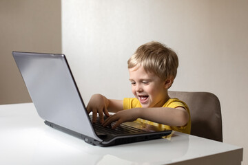 A cute little boy with blond hair sits at a table with a laptop. The boy smiles, he's having fun. He's doing the job.