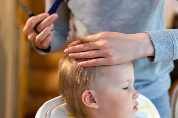 Close-up young adult caucasian mother making haircut fot cute adotrable son toddler boy at home due...