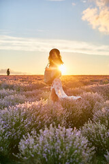 Fototapeta na wymiar Enjoying summer sunset. Young beautiful woman in a straw hat and dress holding basket with lavender flowers and smiling while standing in lavender field at sunset