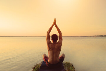 Fit muscular man doing yoga by the water sitting in a lotus pose with hands reaching up the sky...