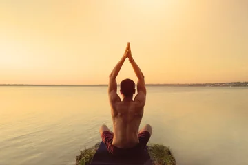 Schilderijen op glas Fit muscular man doing yoga by the water sitting in a lotus pose with hands reaching up the sky namaste. Orange sunlight sunset tranquility at lake. Male back defined muscles. Healthy spine workout © Olivia