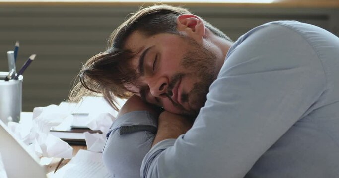 Exhausted lazy young bearded businessman felt asleep at workplace. Overwhelmed stressed male worker employee feeling tired, sleeping on table with paper documents, working hard overnight at home.