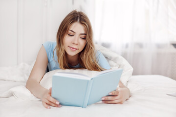 Beautiful girl in blue pajamas reading a book, lying on the bed in the bedroom. Training, education, Hobbies.