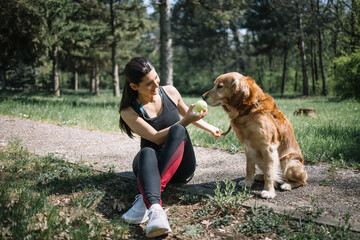 Pretty woman sitting on ground with dog and holding ball. Young woman and her retriever dog playing with tennis ball in park.