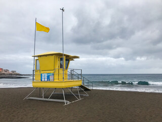 Fototapeta na wymiar View of lifeguard tower on beach during windy sunny day with sea view and unique beach which has black sand on tower flutters green and yellow flag.