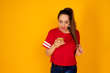 Portrait of positive cheerful girl  with open mouth looks at phone in red t-shirt at  isolated over yellow color background