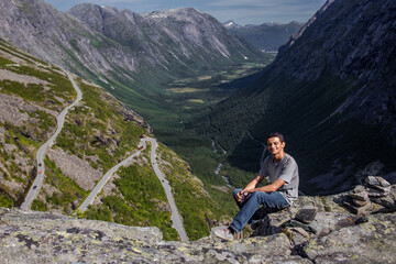 Naklejka premium Trip to Norway. Young hiker sits alone over a cliff and smiles on background of Trollstigen road and green valley with high rocky mountains in bright summer day