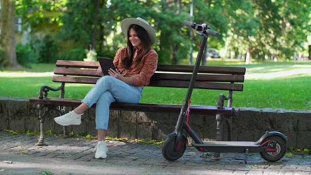 A young woman with a hat is sitting on a park bench and scrolling on the tablet in her hands. An electric scooter is parked next to her, while trees and green colours predominate in the background.