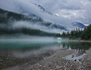 This beautiful landscape is a mountain lake with evergreen trees on the mountain and cloudy skies, with heavy patterns of fog wisping through.  Taken at Ross Lake, Washington, America.