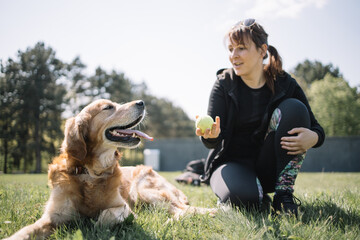 Dog and girl playing with ball outdoor. Blurred woman holding tennis ball while squatting next to retriever dog which is lying on ground in playground.