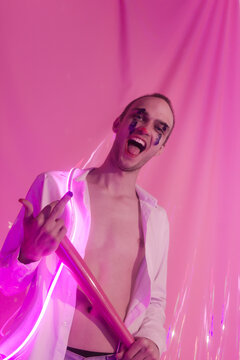 Portrait of Young Handsome emotional man in an unbuttoned white shirt with pink bat. Male in the image of a crazy clown like a joker. Neon pink room. Reflect of neon. Selective focus.