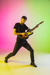 Fototapeta premium Close up young caucasian musician playing bass guitar on gradient studio background in neon. Concept of music, hobby, festival. Colorful portrait of modern artist. Inspired, impressive improvising.