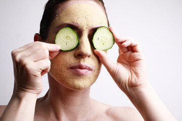 Portrait of young woman with a face mask on holding a cucumber slices in front of one eye. Beauty...