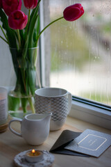 Still life coffee at home, a cozy coffee, tulips in a vase. Coffee on the windowsill, comfort at home. Milk jug  mug, candlestick, envelope and postcard on wooden desk. Spring content