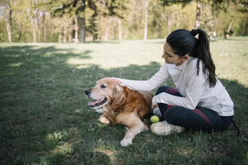 Girl caring dog which is lying on meadow. Brunette girl in sportswear sitting on ground while petting retriever dog in woods.