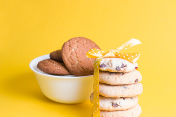 cookies with chocolate pieces and lingonberries tied with a yellow peeling ribbon with a bow on a yellow background copy space, oatmeal cookies in a white bowl in the background