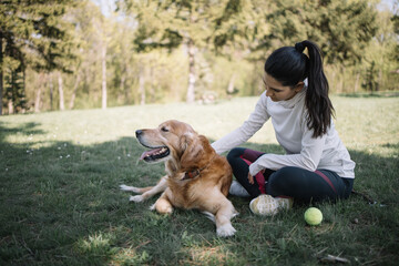 Dog and girl sitting on meadow with tennis ball. Woman in sportswear sitting next to her dog while resting on grass in forest.