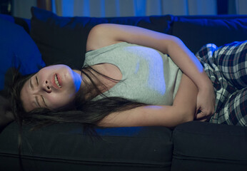 painful menstruation cycle - young beautiful and stressed Asian Korean woman suffering period pain lying on living room sofa couch stressed holding her belly