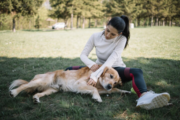 Brunette woman petting dog which is lying on meadow. Sport girl petting her dog while sitting on ground in park in sunny day.