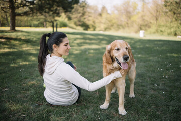 Brunette woman with eyes closed petting dog in nature. Dog standing on meadow and woman petting him while sitting on ground in forest.