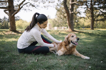 Girl and dog lying on ground in park. Brunette woman with sportswear petting dog while resting on field with trees.