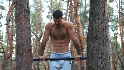 Strong and muscular man doing push ups on horizontal bar in forest. Hardy sportsman working out at nature. Athletic guy with emotional face training outdoor. Concept of sport and active lifestyle