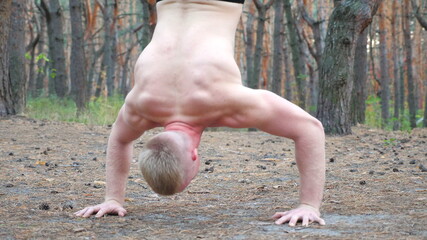 Young muscular man doing push upsin handstand at forest. Hardy and strong guy doing stunts at wood. Close up torso of athlete showing gymnastic exercises outdoor. Sportive and healthy lifestyle