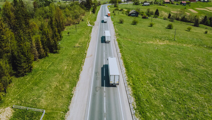 Aerial view to the convoy of trucks going through mountains with some trees and beautiful fields at the side. 