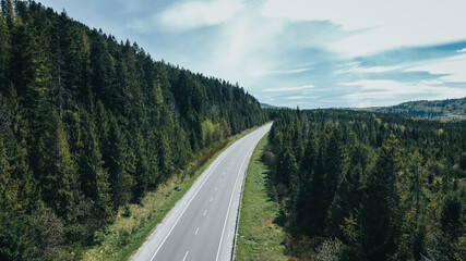 Aerial view to the mountainian road going through the forest with some trees on the sides and blue sky above. 