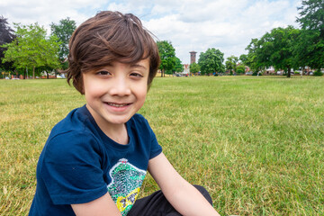 A young boy sits on the floor in the park, smiles and poses for a  portrait.