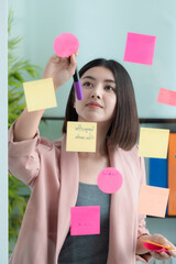 Young Asian businesswoman write tasks creative ideas and planning on sticky post it note on glass wall.