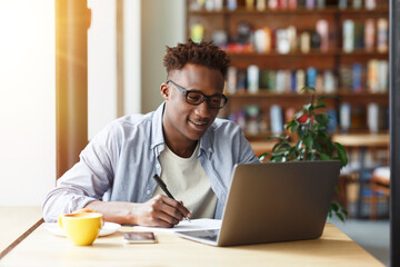 Obraz premium Millennial African American student with laptop taking notes during breakfast in cafe