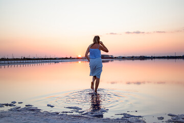 Fototapeta premium Rear view of the woman back look to the purple sunrise in water on summer. Woman walking on the shore of the salt beach at pink lake. vacation lifestyle near ocean