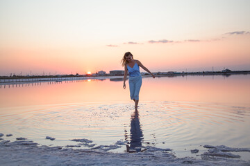 Fototapeta premium Beautiful sensual woman walking and look to the purple sunrise in water on summer. Woman sit on the shore of the salt beach at pink lake. vacation lifestyle near ocean