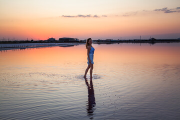 Fototapeta premium View of the woman back silhouette look to the purple sunrise in water on summer. Woman walking on the shore of the salt beach at pink lake. vacation lifestyle near ocean