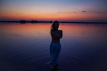 Fototapeta premium The silhouette of woman look to the purple sunrise in water on summer. Woman sit on the shore of the salt beach at pink lake. vacation lifestyle near ocean