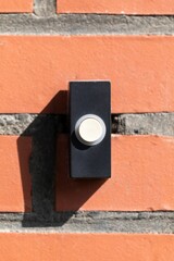 A close up straight portrait of a black and white old plastic doorbell on a red brick wall. The device is ready to be used for someone to announce that they have arived by pressing it and ringing it.