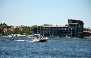 Fototapeta na wymiar A view of a big multi-storey hotel located next to the edge of a vast river or lake with a single white boat travelling along the reservoir dynamically on a cloudless summer day in Poland